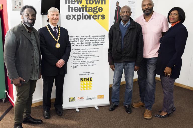 Picture of Mayor Sam Crooks with celebrated Black Pioneers in Milton Keynes standing in front of their exhibition and the New Town Heritage Explorers Logo