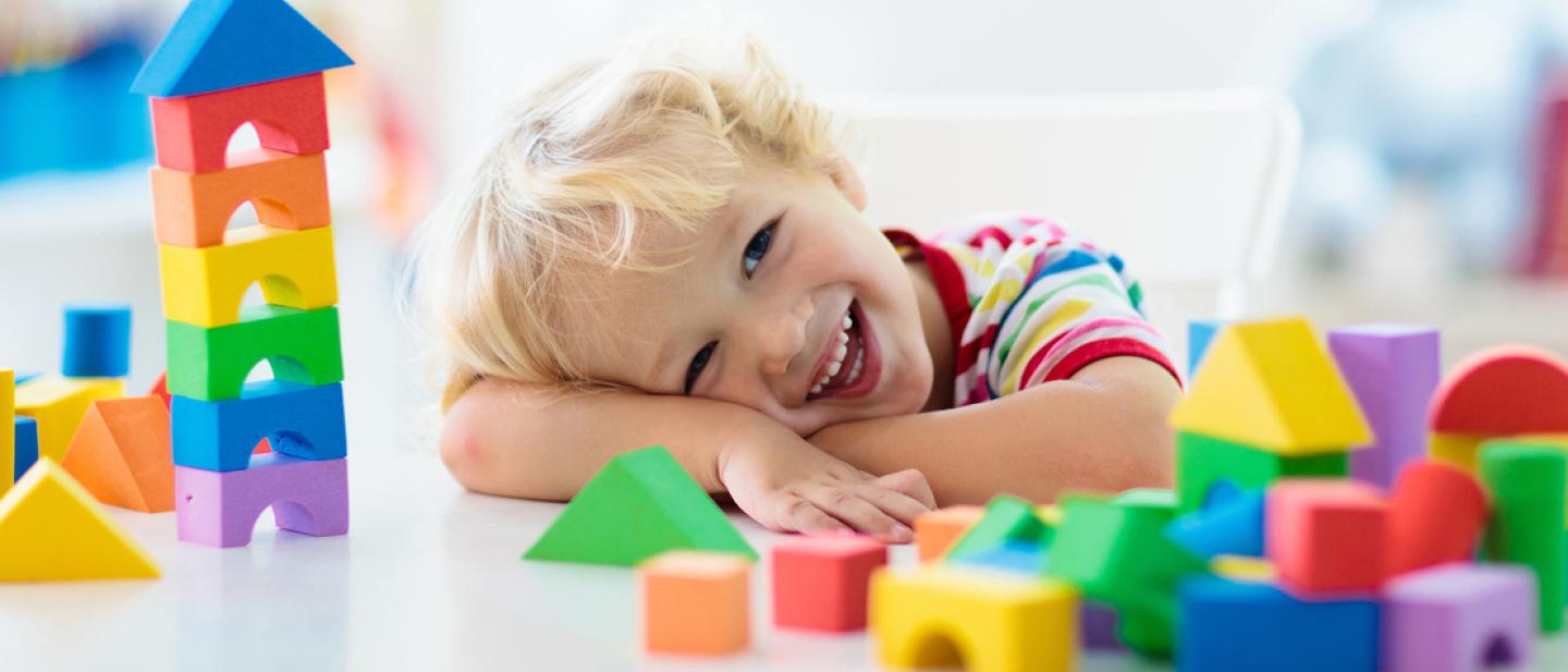 A toddler smiling at the camera while resting his head on a table filled with toy building blocks.