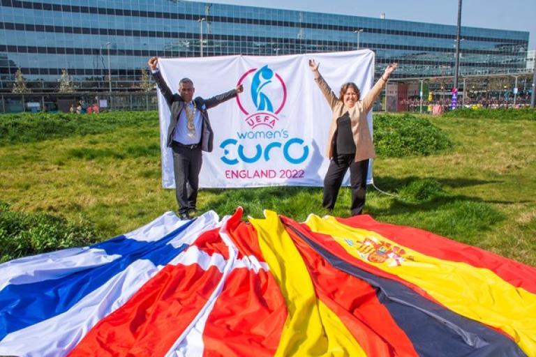 Mayor of Milton Keynes, Councillor Mohammed Khan and Cabinet Member for Sport Councillor Jane Carr holding the UEFA Women's Euro 2022 flag, standing in front of the national flags of Germany, Denmark, Spain, and Finland.