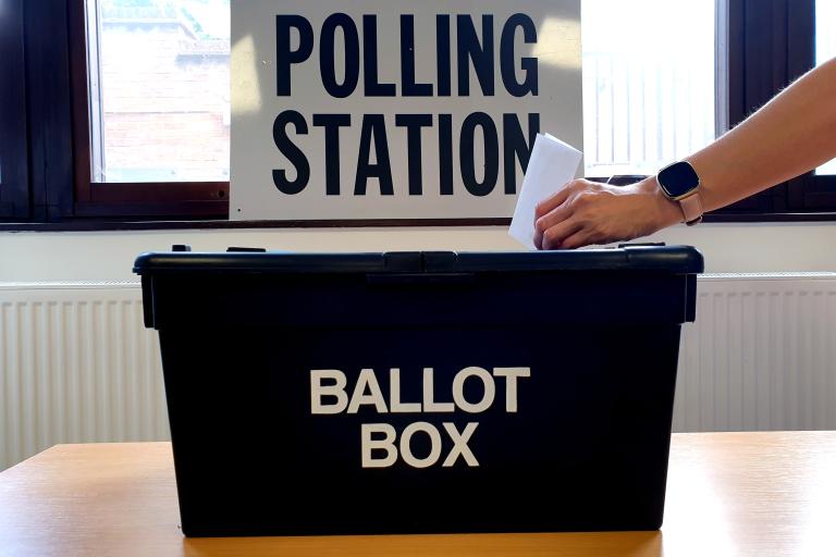 Hand placing ballot into ballot box with Polling station sign in the background