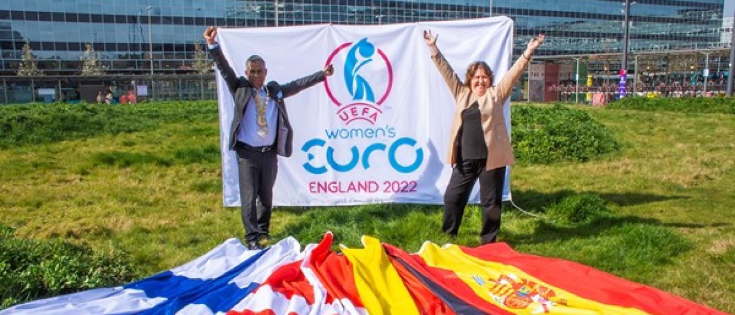 Mayor of Milton Keynes, Councillor Mohammed Khan and Cabinet Member for Sport Councillor Jane Carr holding the UEFA Women's Euro 2022 flag, standing in front of the national flags of Germany, Denmark, Spain, and Finland.