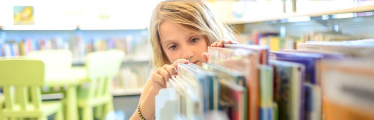 A young girl looking at books on a library shelf.