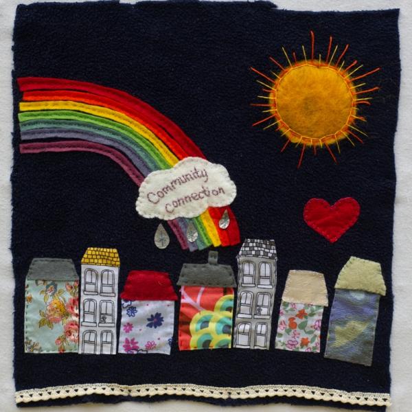 Quilt square with a row of houses made out of fabric with a rainbox, sun and heart in the sky. Text on a cloud reads: Community Connection