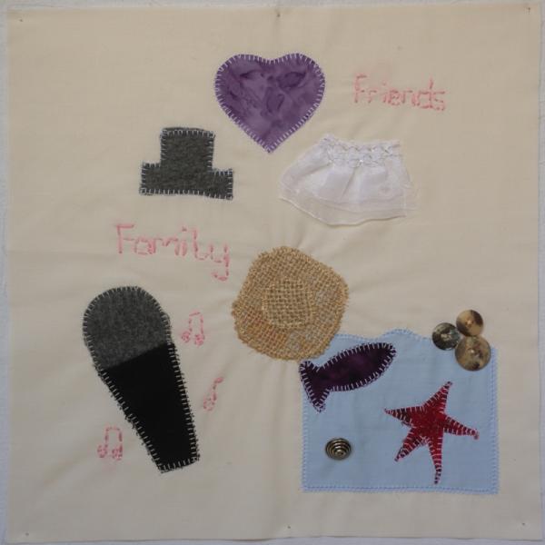 Quilt square with objects sewn on to represent a microphone, music, a heart, wedding dress, beach towel, hat, fish and starfish. Text reads: Friends Family.